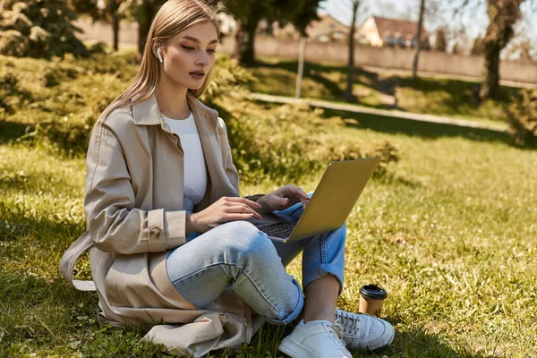 Blonde young woman in earphones and trench coat using laptop while sitting on grass near paper cup — Stock Photo