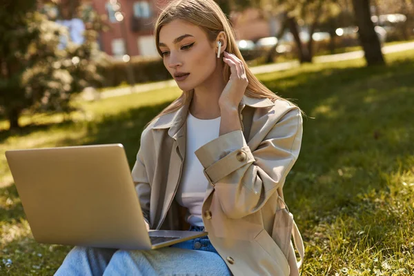 Blonde young woman in wireless earphones and trench coat using laptop while sitting on grass — Stock Photo
