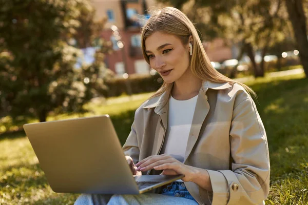 Приваблива молода жінка в бездротових навушниках і траншеї пальто, використовуючи ноутбук, сидячи на траві — стокове фото