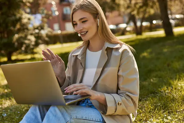 Mujer joven feliz en auriculares inalámbricos y gabardina usando el ordenador portátil mientras tiene videollamada - foto de stock