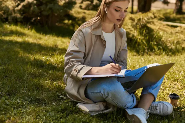 Blonde woman in earphones and trench coat doing online research on laptop while writing in notebook — Stock Photo