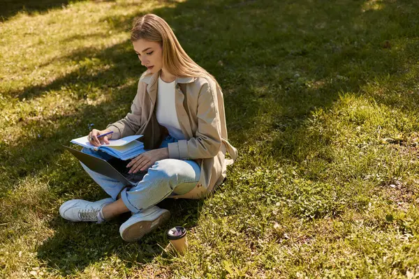 Mujer rubia en jeans y gabardina haciendo investigación en línea en el ordenador portátil, mientras que la escritura en cuaderno - foto de stock
