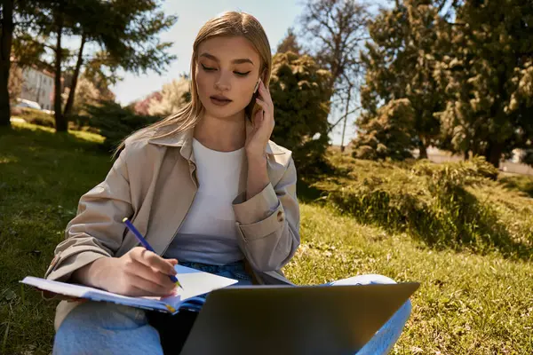 Блондинка в траншеї пише в блокноті, використовуючи ноутбук і сидячи на газоні в парку — стокове фото