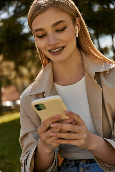 Mujer rubia feliz en auriculares inalámbricos y gabardina beige usando su teléfono inteligente en el parque - foto de stock