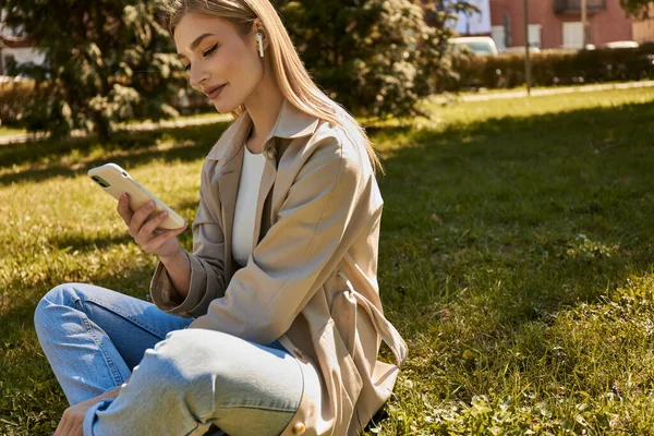 Happy blonde woman in wireless earphones and trench coat using her smartphone and sitting on lawn — Stock Photo