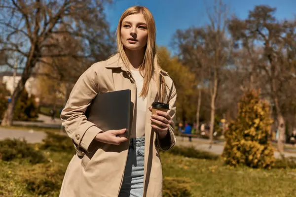 Hübsche blonde Frau im schicken Trenchcoat mit Pappbecher mit Kaffee und Laptop im Park — Stockfoto