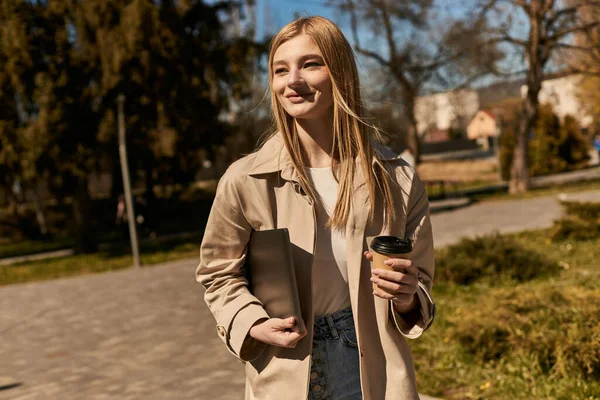 Happy blonde woman in stylish trench coat holding paper cup with coffee and laptop in park — Stock Photo