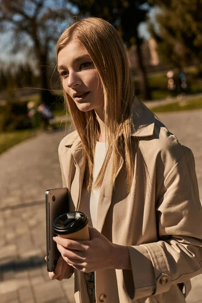 Junge blonde Frau im beigen Trenchcoat hält Pappbecher mit Kaffee und Laptop in der Hand und geht im Park spazieren — Stockfoto