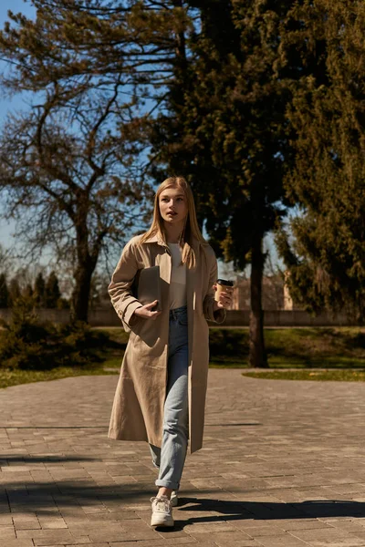 Young blonde woman in trench coat holding paper cup with to go coffee and laptop and walking in park — Stock Photo