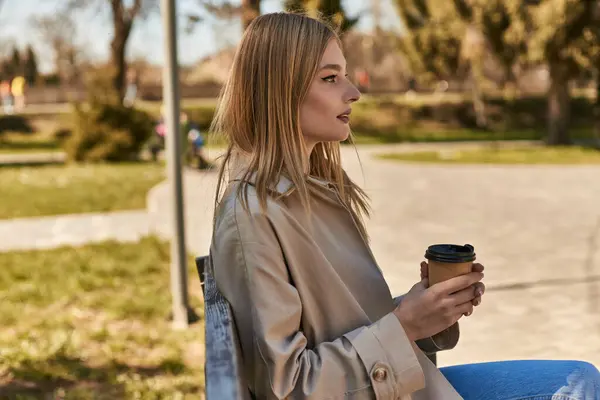 Jeune femme blonde en trench coat tenant tasse en papier avec café à emporter, assise sur un banc dans le parc — Photo de stock