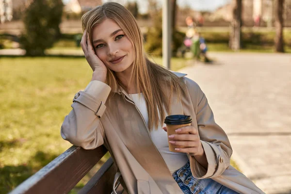 Jovem mulher feliz em casaco de trincheira segurando copo de papel com café takeaway, sentado no banco no parque — Fotografia de Stock