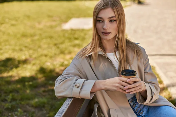 Joven rubia en gabardina sosteniendo taza de papel con café y sentada en el banco en el parque - foto de stock