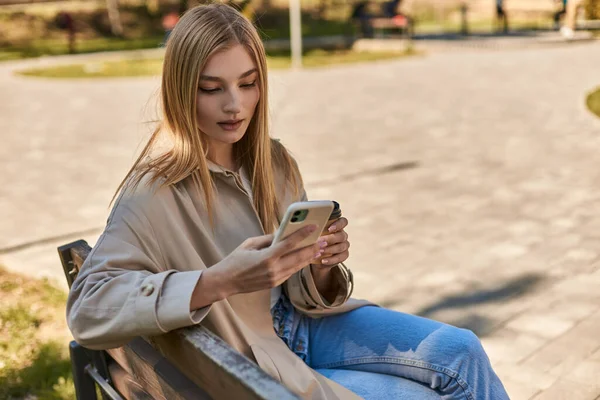Jeune femme en trench coat tenant tasse en papier avec café et en utilisant smartphone tout en étant assis sur le banc — Photo de stock