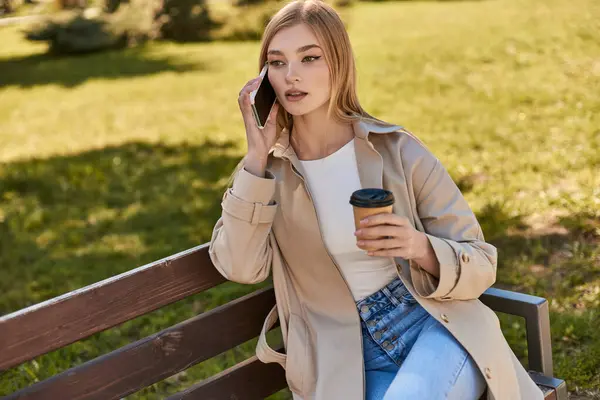 Young woman in trench coat holding takeaway coffee and talking on smartphone while sitting on bench — Stock Photo