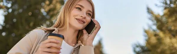 Estandarte de la mujer feliz en gabardina celebración de café para llevar y hablar en el teléfono inteligente al aire libre - foto de stock