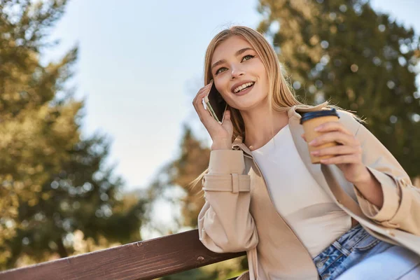 Happy woman in trench coat holding coffee to go and talking on smartphone while sitting on bench — Stock Photo