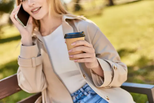 Frau im Trenchcoat hält Coffee to go in der Hand und spricht auf der Bank mit dem Smartphone — Stockfoto