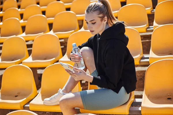 Спортивная женщина с хвостиком, держащая бутылку воды и используя смартфон после тренировки на стадионе — стоковое фото