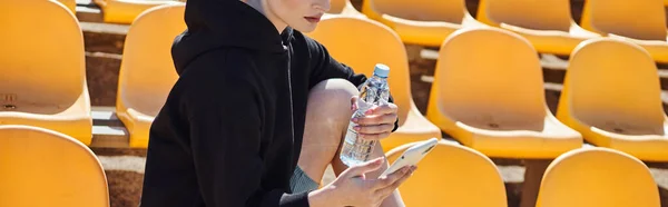 Cropped sporty woman holding bottle of water and using smartphone after workout in stadium, banner — Stock Photo