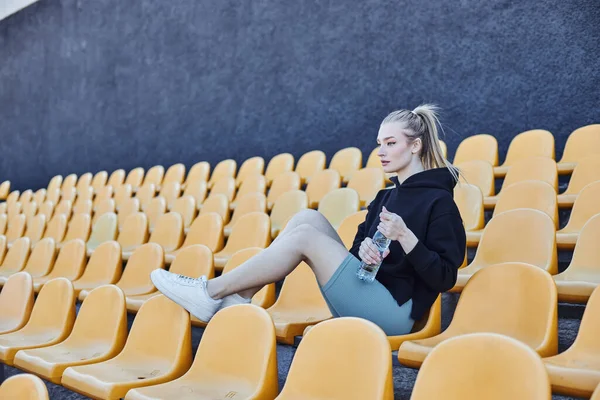 Sportswoman with ponytail holding bottle of water and sitting on stadium chair after workout — Stock Photo