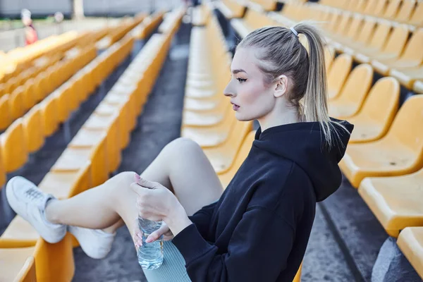 Блондинка с хвостиком держит бутылку воды и сидит на стуле стадиона после тренировки — стоковое фото