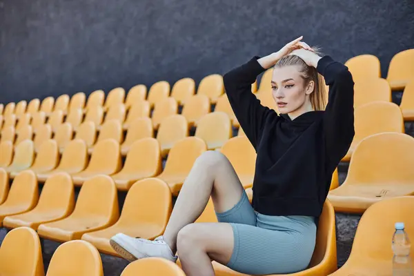 Blonde Sportlerin macht ihren Pferdeschwanz und sitzt nach dem Training auf einem Plastikstuhl im Stadion — Stockfoto