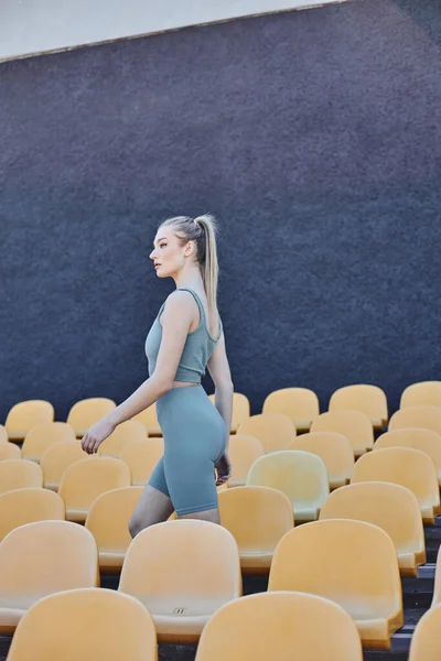 Atlética e loira esportista em activewear passando por cadeiras de estádio amarelo, moda esportiva — Fotografia de Stock