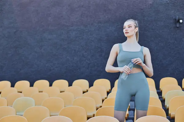 Athletische und blonde Sportlerin in Activwear mit Wasserflasche in der Nähe gelber Stadionstühle — Stockfoto