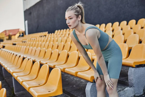 Pretty and blonde sportswoman in activewear exercising near yellow stadium chairs, physical activity — Stock Photo