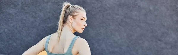 Pretty and blonde sportswoman in activewear posing next to grey wall outdoors, horizontal banner — Stock Photo