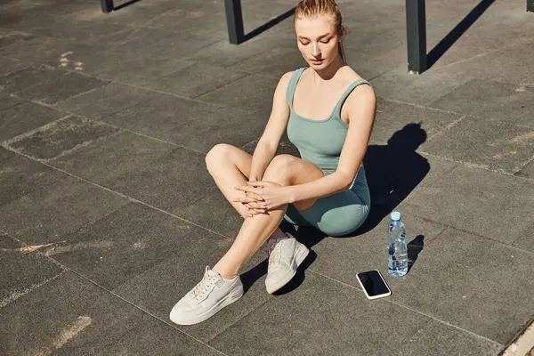 Blonde sportswoman in tight activewear sitting next to bottle of water and smartphone on floor — Stock Photo