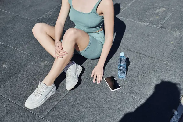 Cropped sportswoman in tight activewear sitting next to bottle of water and smartphone on floor — Stock Photo
