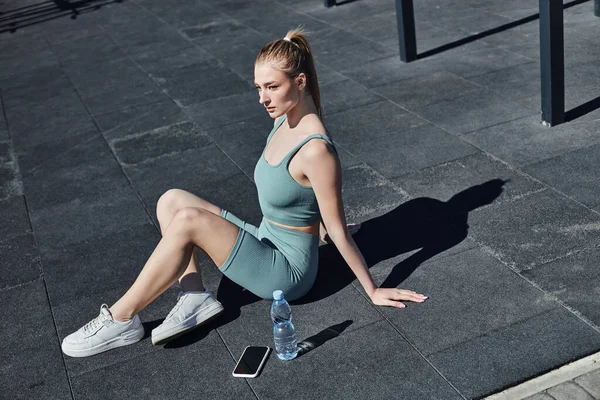 Young fit woman in tight activewear sitting next to bottle of water and smartphone after workout — Stock Photo