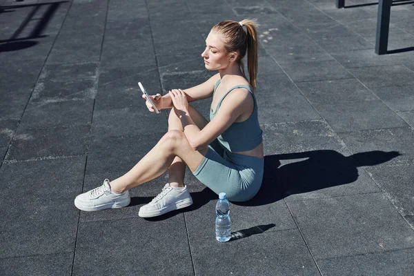 Mujer en forma de ropa deportiva apretado sentado junto a la botella de agua y el uso de teléfono inteligente después del entrenamiento - foto de stock