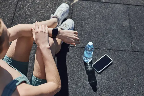 Top view of fit woman in activewear checking fitness tracker next to smartphone and water bottle — Stock Photo