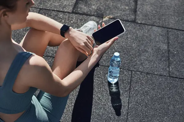 Top view of fit woman in activewear with fitness tracker on wrist using smartphone near water bottle — Stock Photo