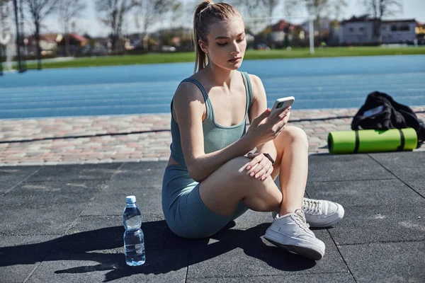 Young fit woman in activewear using smartphone and sitting near water bottle, health and fitness — Stock Photo