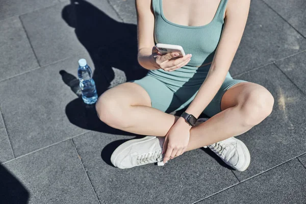 Обрізана підходить жінка в активному одязі за допомогою смартфона і сидить біля пляшки води, здоров'я і фітнесу — стокове фото