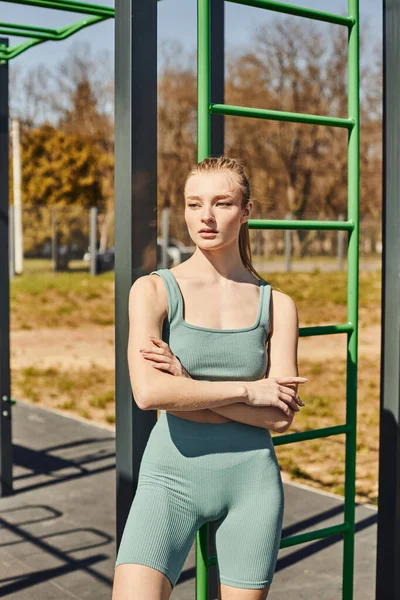 Young blonde woman in tight activewear standing with crossed arms near vertical ladder outside — Stock Photo
