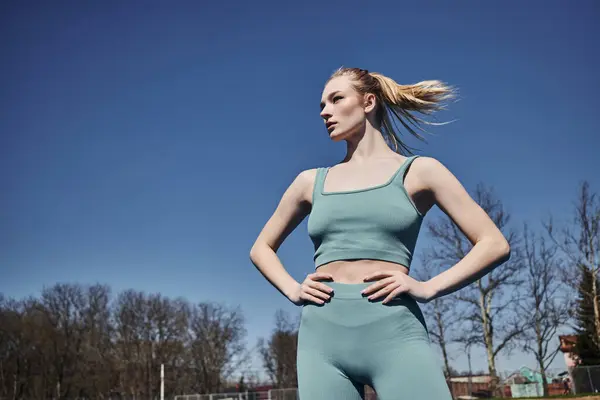 Low angle view of blonde sportswoman in activewear standing with hands on hips and looking away — Stock Photo