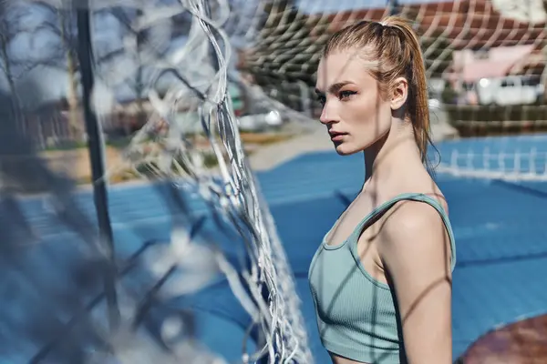 Blonde and fit woman with ponytail standing in activewear near net after working out outdoors — Stock Photo