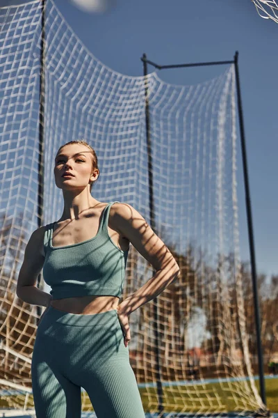 Молодая женщина с хвостиком, позирующая в спортивной одежде возле сетки во время тренировки на открытом воздухе, солнечная погода — стоковое фото