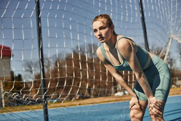 Спортивная молодая женщина с хвостиком в активной одежде отдыхает во время тренировки на открытом воздухе возле сети — стоковое фото
