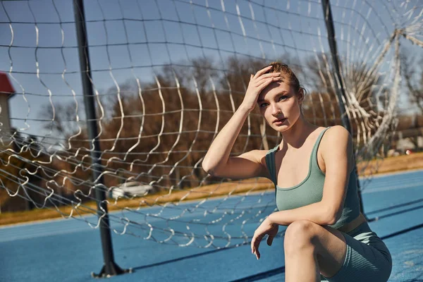 Mujer joven rubia con cola de caballo en ropa deportiva descansando mientras está sentada en las horcas después del entrenamiento - foto de stock