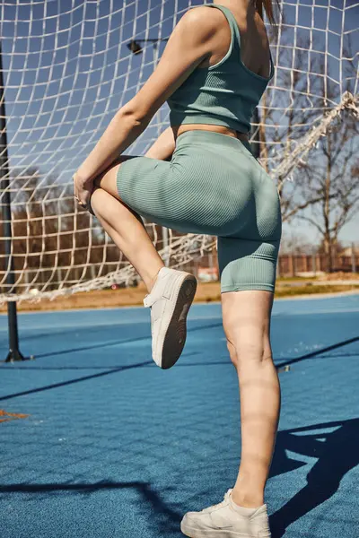 Обрезанные молодая спортсменка упражнения в активной одежде и кроссовки возле сети на открытом воздухе, городской фитнес — стоковое фото
