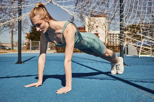 Blonde young sportswoman with ponytail exercising in activewear near net outdoors, doing press ups — Stock Photo
