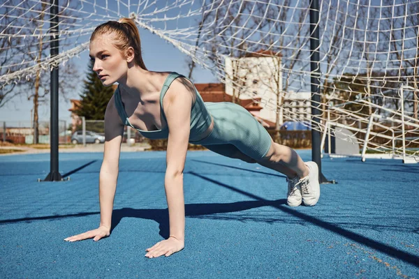Pretty young sportswoman with ponytail exercising in activewear near net outdoors, doing press ups — Stock Photo