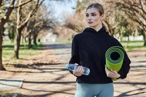 Loira e apto jovem mulher em preto desportivo capuz segurando tapete de fitness e garrafa de água no parque — Fotografia de Stock