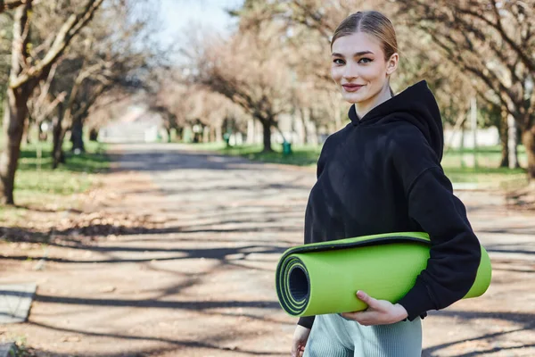 Blonde and fit young woman in black sporty hoodie holding fitness mat after working out in park — Stock Photo