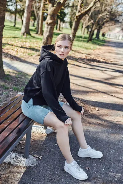 Blonde and fit young woman in black sporty hoodie and cycling shorts sitting on wooden bench in park — Stock Photo